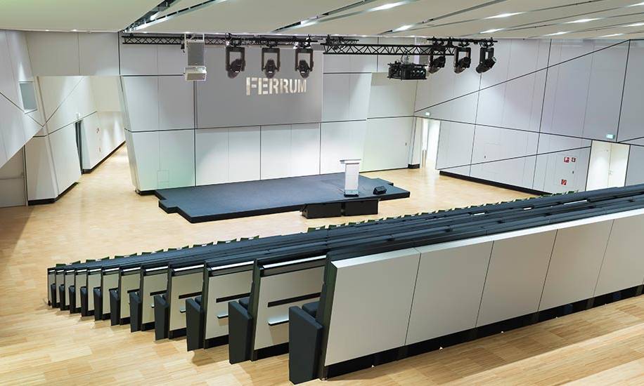 Blick von der linken obersten Stelle in den Konferenzraum ferrum. Im Raum sind standardmäßig ansteigende Sitzreihen mit eingebaut. In der Bildmitte erkennt man das dunkel gehaltene Podium mit Sprechpult und die dahinterstehende weißen Wand, die als Leinwand nutzbar ist. Die weißen Wände werden auf der linken Seite mit grünen Strahlern angeleuchtet.